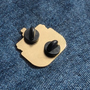 May include: Gold-colored metal pin with two black metal clasps on the back. The pin is shaped like a cartoon character with a large head and a small body.