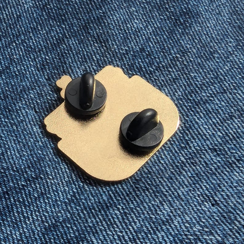 May include: Gold-colored metal pin with two black metal clasps on the back. The pin is shaped like a cartoon character with a large head and a small body.