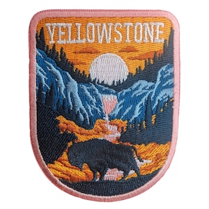 May include: Embroidered patch depicting a Yellowstone National Park scene. The design shows a bison, mountains, a waterfall, and a sunset, with the word "YELLOWSTONE" embroidered above. The patch has a pink border.