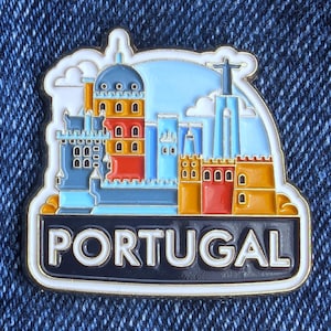 Puede incluir: Un colorido pin de esmalte con una representación estilizada de los monumentos de Portugal. El pin muestra un paisaje urbano con edificios en azul, naranja y rojo, con la palabra "PORTUGAL" en letras blancas sobre una pancarta negra.