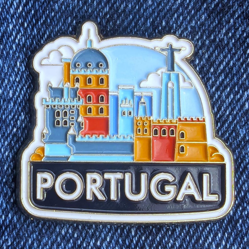 Op de afbeelding: Een kleurrijke emaille pin met een gestileerde afbeelding van de bezienswaardigheden van Portugal. De pin toont een stadsgezicht met gebouwen in blauw, oranje en rood, met het woord "PORTUGAL" in witte letters op een zwarte banner.