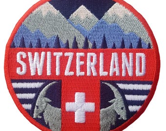 Può includere: Toppa rotonda ricamata con la scritta "SWITZERLAND" in bianco. Presenta uno sfondo rosso con una croce bianca, montagne blu e bianche e due stambecchi grigi. Il bordo è rosso.
