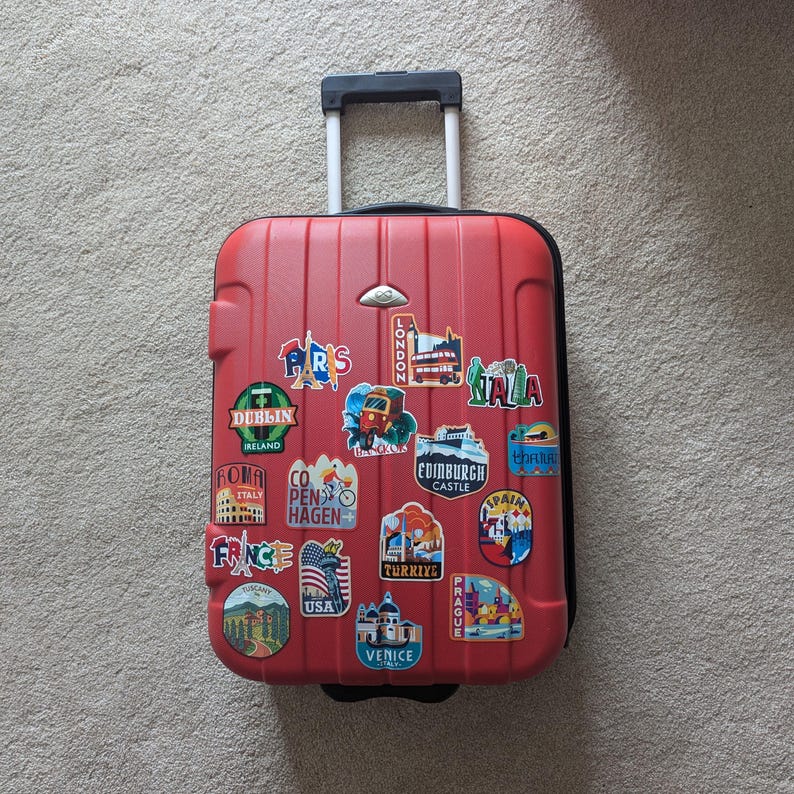 May include: Red suitcase with a handle and wheels covered in travel stickers from various cities around the world, including Paris, London, Dublin, Rome, Copenhagen, Edinburgh, Spain, Turkey, Prague, Venice, and USA.