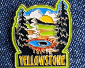 May include: A colorful Yellowstone National Park enamel pin. The pin features a landscape with a yellow sun, blue sky, green and brown terrain, and black trees. The word "YELLOWSTONE" is displayed in yellow letters.