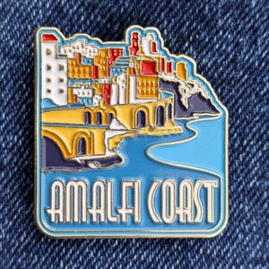 May include: A colorful enamel pin featuring the Amalfi Coast. The pin depicts a coastal town with buildings in shades of white, yellow, red, and orange, set against a blue sea and sky. The words "AMALFI COAST" are displayed in a bold, stylized font.