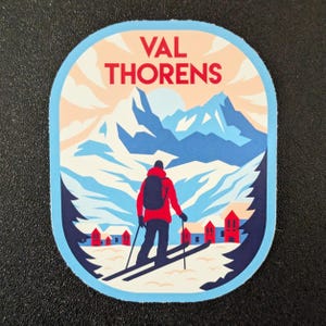 Könnte beinhalten: Ovaler Aufkleber mit einem Skifahrer in einer roten Jacke und einem schwarzen Rucksack, der auf rote Gebäude zufährt. Der Hintergrund zeigt blaue Berge und den Text "VAL THORENS" in Rot.