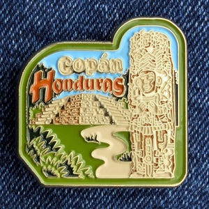 May include: A rectangular Copán Honduras souvenir pin with a green border. The pin features a light blue sky, a brown pyramid, and a detailed carving of a figure. The words "Copán Honduras" are in orange and yellow.