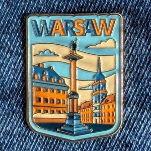 May include: A rectangular enamel pin with the word "WARSAW" in orange and blue. The pin shows a cityscape with buildings, a tall column, and a blue sky. The pin is set against a denim background.