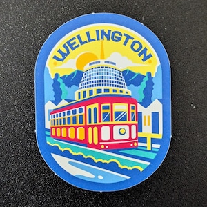 May include: A blue sticker with the word "WELLINGTON" in yellow. The sticker features a red and yellow trolley car, a domed building, and a yellow sun over a mountain range. The sticker is an oval shape.