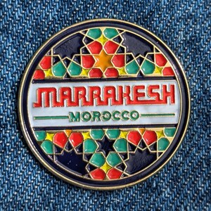 Puede incluir: Un pin redondo y decorativo con borde dorado. El pin presenta la palabra "MARRAKESH" en letras rojas sobre "MOROCCO" en verde. El diseño incluye patrones geométricos en rojo, verde, amarillo y naranja sobre un fondo azul oscuro.