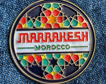 May include: A round, decorative pin with a gold border. The pin features the word "MARRAKESH" in red letters above "MOROCCO" in green. The design includes geometric patterns in red, green, yellow, and orange against a dark blue background.