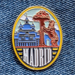 Könnte beinhalten: Ovale Emaille-Anstecknadel mit einem blauen Gebäude, einem roten Baum und einem Bären. Das Wort "MADRID" ist in weißen Blockbuchstaben am unteren Rand vor einem gelb-blauen Hintergrund dargestellt. Die Anstecknadel befindet sich vor einem Jeans-Hintergrund.
