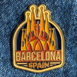 Puede incluir: Un pin de esmalte dorado con la Sagrada Familia de Barcelona, España. El pin tiene un fondo azul marino con las palabras "BARCELONA" y "SPAIN" en letras naranjas y doradas. El diseño arquitectónico es en tonos naranja y amarillo.
