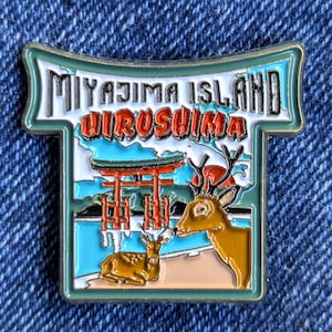 Puede incluir: Pin esmaltado con la isla de Miyajima, Hiroshima. El diseño incluye una puerta torii roja, ciervos y el texto "Miyajima Island Hiroshima". El pin tiene un borde turquesa y blanco.