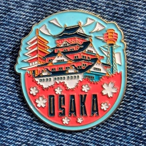 Puede incluir: Un colorido pin de esmalte con monumentos de Osaka. El pin tiene un borde azul claro, una pagoda roja y blanca, un castillo de varios niveles y una torre. La palabra "OSAKA" se muestra en negrita, rodeada de acentos florales blancos.