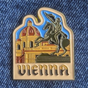Könnte beinhalten: Rechteckiger Emaille-Pin mit einer Szene aus Wien, Österreich. Der Pin zeigt ein Gebäude mit roter Kuppel, einen Reiter auf einem Pferd und das Wort "VIENNA" in einer dekorativen Schriftart. Der Pin hat einen goldenen Rand und ist vor blauem Himmel platziert.