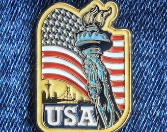 May include: A gold-coloured pin depicting the Statue of Liberty holding a torch, set against the American flag. The pin includes the word "USA" and a city skyline silhouette. The pin is rectangular with a gold border.