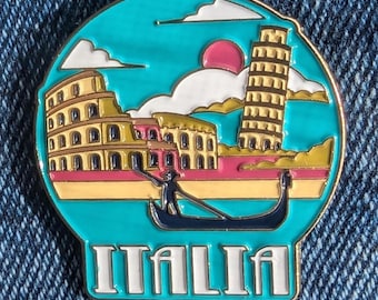 May include: A round, teal-colored enamel pin featuring Italian landmarks. The design includes the Colosseum, the Leaning Tower of Pisa, and a gondola. The word "ITALIA" is displayed in white block letters at the bottom, with a gold border.