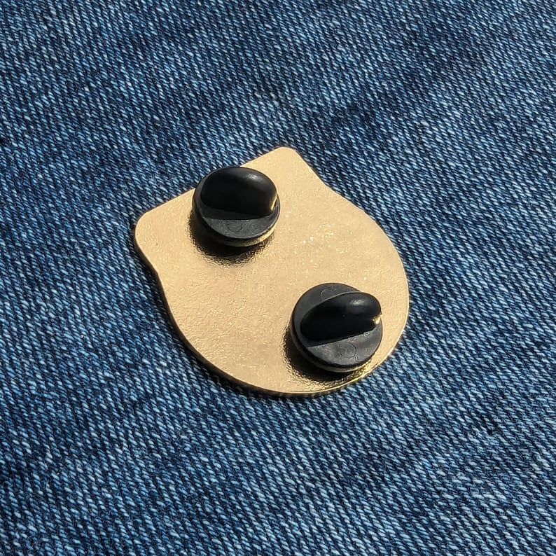May include: Gold-colored metal pin with two black pin backs. The pin is shaped like a rounded rectangle.