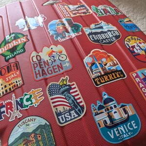 May include: Red suitcase covered in colorful travel stickers with city names and landmarks, including Dublin, Ireland, Roma, Italy, London, Copenhagen, Edinburgh Castle, Spain, Turkey, Prague, France, Tuscany, USA, and Venice, Italy.