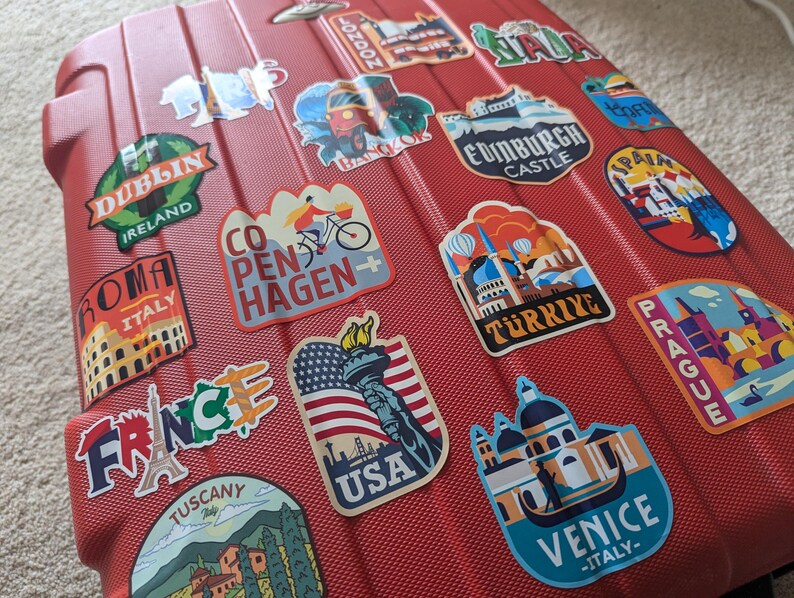 May include: Red suitcase covered in colorful travel stickers with city names and landmarks, including Dublin, Ireland, Roma, Italy, London, Copenhagen, Edinburgh Castle, Spain, Turkey, Prague, France, Tuscany, USA, and Venice, Italy.