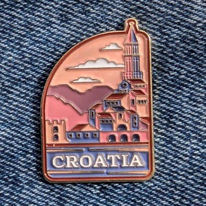May include: A colorful enamel pin featuring a Croatian cityscape with a tower, buildings, and mountains under a sunset sky. The word "CROATIA" is displayed at the bottom of the pin, which has a gold border.