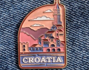 May include: A colorful enamel pin featuring a Croatian cityscape with a tower, buildings, and mountains under a sunset sky. The word "CROATIA" is displayed at the bottom of the pin, which has a gold border.