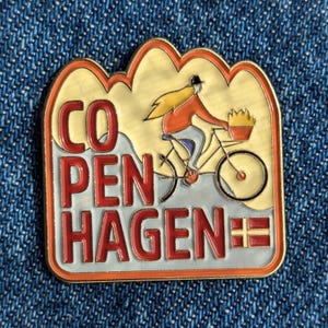 Puede incluir: Pin esmaltado con la palabra "Copenhagen" en rojo. El pin representa a una persona montando en bicicleta con una cesta y la bandera danesa. El pin tiene un fondo amarillo y crema.