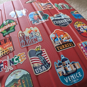 May include: Red suitcase covered in colorful travel stickers with city names and landmarks, including Dublin, Ireland, Roma, Italy, London, Copenhagen, Edinburgh Castle, Spain, Turkey, Prague, France, Tuscany, USA, and Venice, Italy.