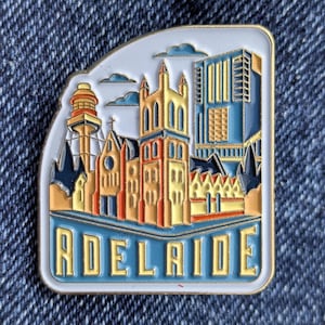 Puede incluir: Un pin rectangular de esmalte con un paisaje urbano estilizado y la palabra "ADELAIDE" en azul y blanco. El pin muestra edificios en tonos amarillos, naranjas y azules, sobre un cielo azul claro, con un borde blanco.