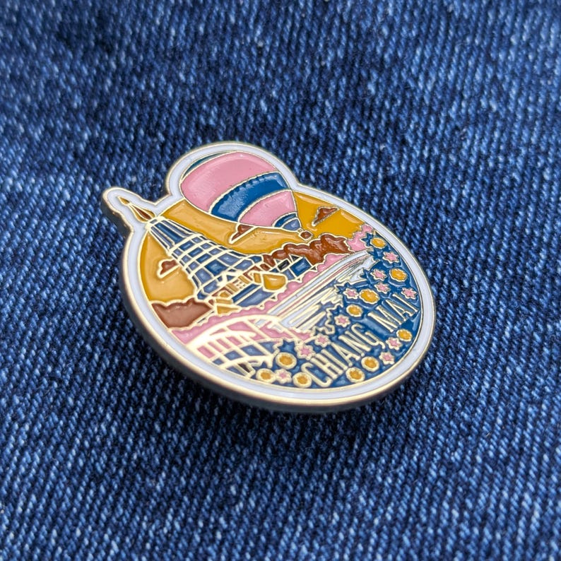 K&ouml;nnte beinhalten: Emaille-Anstecknadel mit einem Chiang Mai-Design. Die Anstecknadel zeigt einen rosa und blauen Hei&szlig;luftballon, eine Pagode und die Worte "Chiang Mai" in wei&szlig;en Buchstaben. Die Anstecknadel hat einen goldenen Rand und ist vor einem blauen Jeanshintergrund platziert.