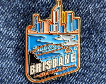 May include: A rectangular enamel pin featuring a Brisbane, Australia cityscape. The pin has a blue river, a bridge, and buildings in shades of blue, orange, and gold. The word "BRISBANE" is in white, and "AUSTRALIA" is in gold.