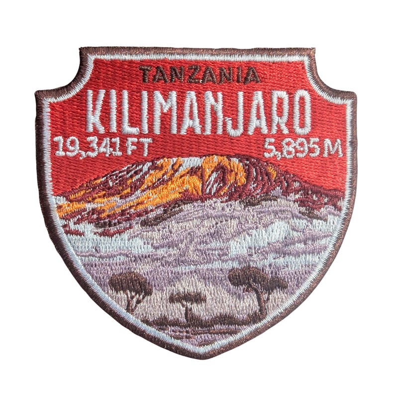 May include: Embroidered patch featuring Mount Kilimanjaro in Tanzania. The red patch has white text that reads "TANZANIA KILIMANJARO" and the elevation in feet and meters. The mountain is depicted in shades of orange, brown, and purple.