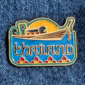 May include: A rectangular enamel pin with a gold border and the word "thailand" in blue. The pin features a boat with a yellow sun in the background. The boat is brown with red and white accents. The pin is on a blue denim background.