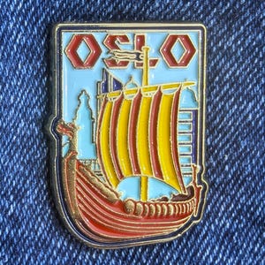 May include: A rectangular pin badge with a Viking ship design. The pin features yellow and red sails, the word "OSLO" in red, and a light blue backdrop. It has a gold border and is set against a blue denim background.