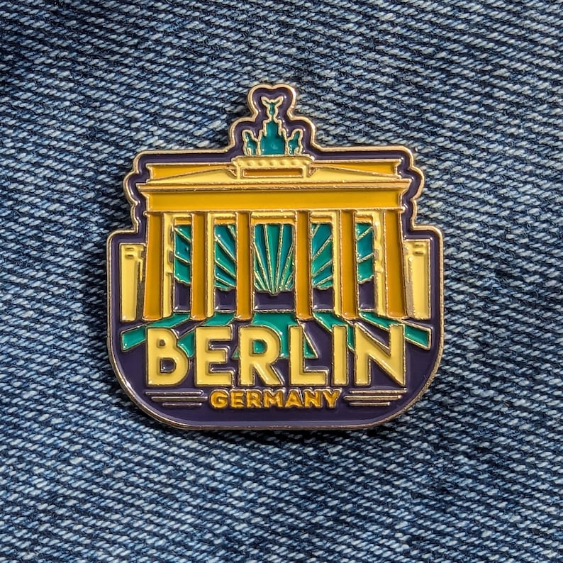 May include: A gold-colored enamel pin featuring a stylized illustration of the Brandenburg Gate in Berlin, Germany. The pin has a purple background with the word "BERLIN" in yellow letters and "GERMANY" in smaller yellow letters.