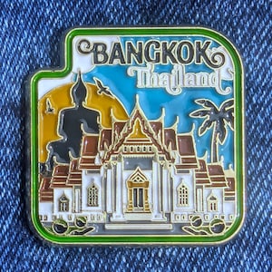 May include: A rectangular Bangkok, Thailand souvenir pin with a green border. The pin features a temple, a Buddha statue, a palm tree, and the words "BANGKOK Thailand" in a blue sky background.