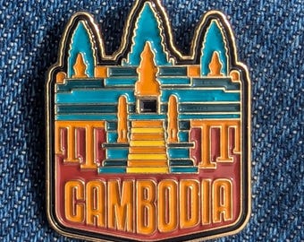May include: A colorful enamel pin featuring a stylized depiction of a temple with the word "CAMBODIA" in orange. The pin has a gold border and is set against a blue denim background. The temple design incorporates teal, orange, and yellow.