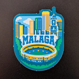 May include: A teal-colored souvenir magnet featuring the city of Malaga. The design includes a skyline with various buildings, a stadium, and the city name in white and blue. The magnet has a unique shape.