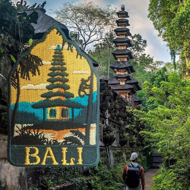 May include: Embroidered patch with a Bali temple design, featuring a multi-tiered structure, palm trees, and the word "BALI" in gold lettering. The patch is set against a lush green backdrop with a person walking on a path.