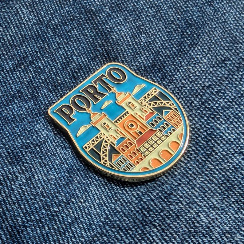 May include: A gold-colored enamel pin with a blue background and a white building with orange accents. The pin features the word "PORTO" in black letters and a depiction of a city skyline.
