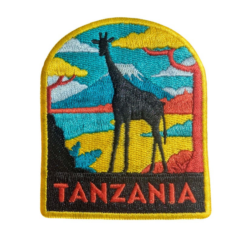 May include: Embroidered patch featuring a black giraffe silhouette against a colorful landscape. The design includes a yellow border, blue sky, and the word "TANZANIA" in red letters. The patch is a travel-themed souvenir.