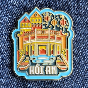 Op de afbeelding: Emaille pin met een kleurrijke illustratie van een Vietnamese architectonische bezienswaardigheid. De pin heeft een lichtblauwe rand en het woord "HOI AN" in witte letters. Het ontwerp bevat oranje, gele en rode tinten.