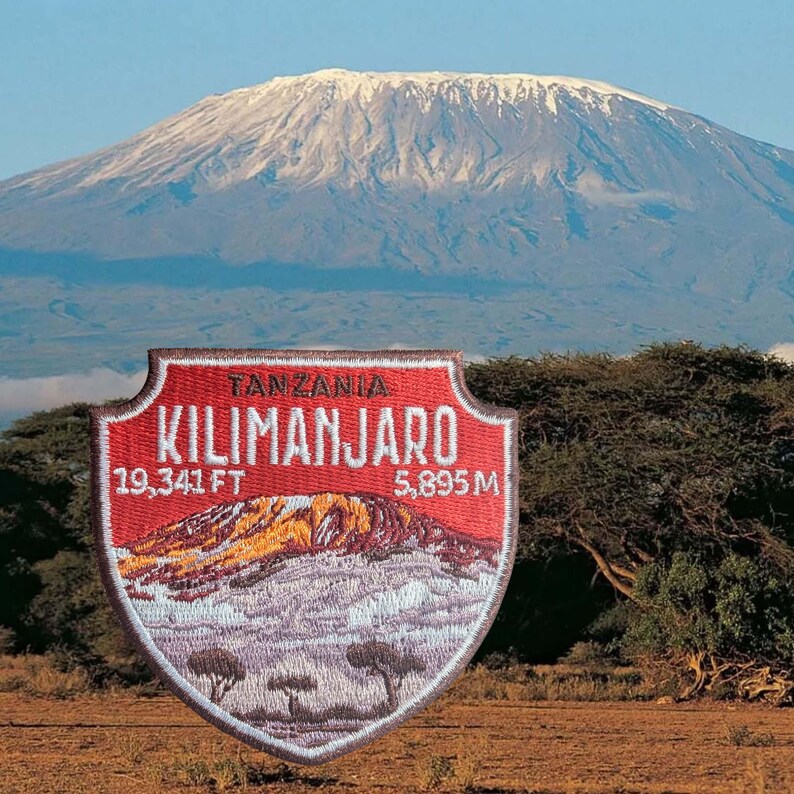 May include: A red and white embroidered patch featuring a mountain landscape and the text "TANZANIA KILIMANJARO". The patch includes the measurements "19,341 FT" and "5,895 M". The background shows a snow-capped mountain.
