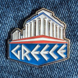 Puede incluir: Pin rectangular con un edificio blanco y azul con columnas, sobre una franja roja y un fondo azul con la palabra "GREECE" en blanco. El pin tiene un borde plateado y está sobre un fondo de mezclilla.