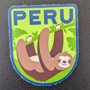Puede incluir: Una pegatina con la palabra "PERU" en azul sobre un fondo verde. Un perezoso marrón cuelga de una rama con hojas verdes. La pegatina tiene un borde azul.