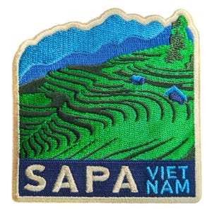 May include: Embroidered patch depicting a green terraced landscape with blue mountains and sky. The patch reads "SAPA VIET NAM" in white and blue on a navy banner. The patch has a white border, suitable for sewing onto clothing or bags.
