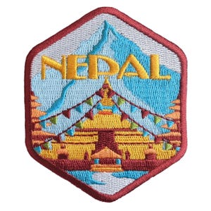 May include: Embroidered patch with a hexagonal shape, displaying the word "NEPAL" in gold. The design includes a mountain range, a pagoda, and colourful flags, all framed by a red border.
