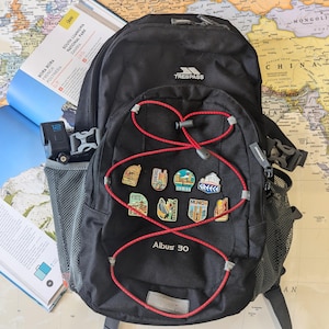K&ouml;nnte beinhalten: Schwarzer Trespass-Rucksack mit roten Akzenten und Reise-Pins. Der Rucksack hat eine Seitentasche aus Mesh und ein rotes Bungee-Cord-Design. Eine Karte und ein Reisef&uuml;hrer sind im Hintergrund zu sehen.