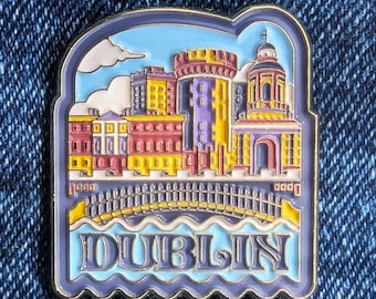 May include: A rectangular enamel pin featuring a colorful depiction of Dublin, Ireland. The pin showcases buildings, a bridge, and the word "DUBLIN" in a stylized font, set against a blue sky and water background. The pin has a gold border.